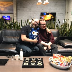 Senior Producer Daniel Smith and composer Gareth Coker seated on a couch with a Ku plush.
